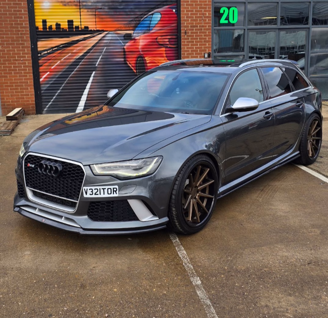 Grey Audi RS6 Avant parked on a concrete surface with bronze alloy wheels, in front of a red brick building and mural featuring a road and car scene