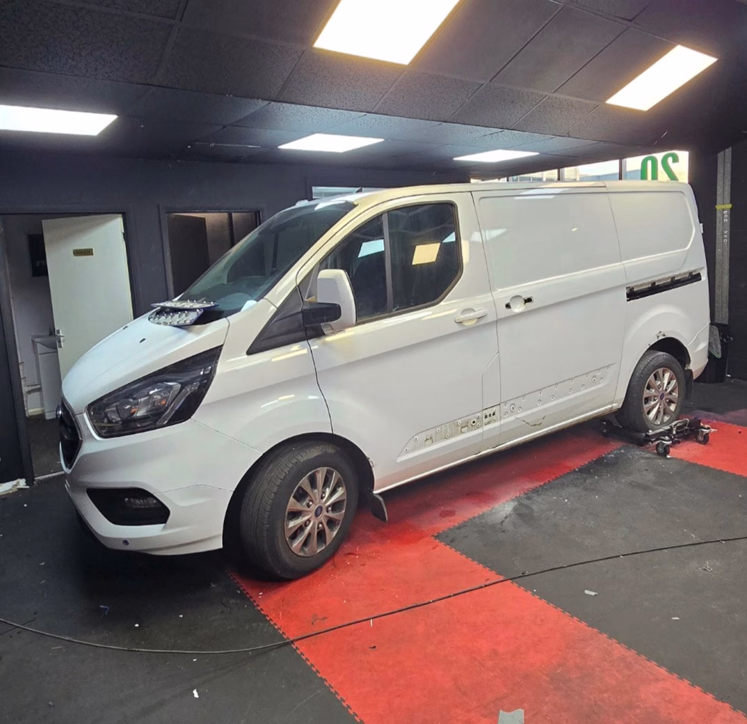 White panel van parked inside a workshop on red and black floor mats with workshop lighting above.