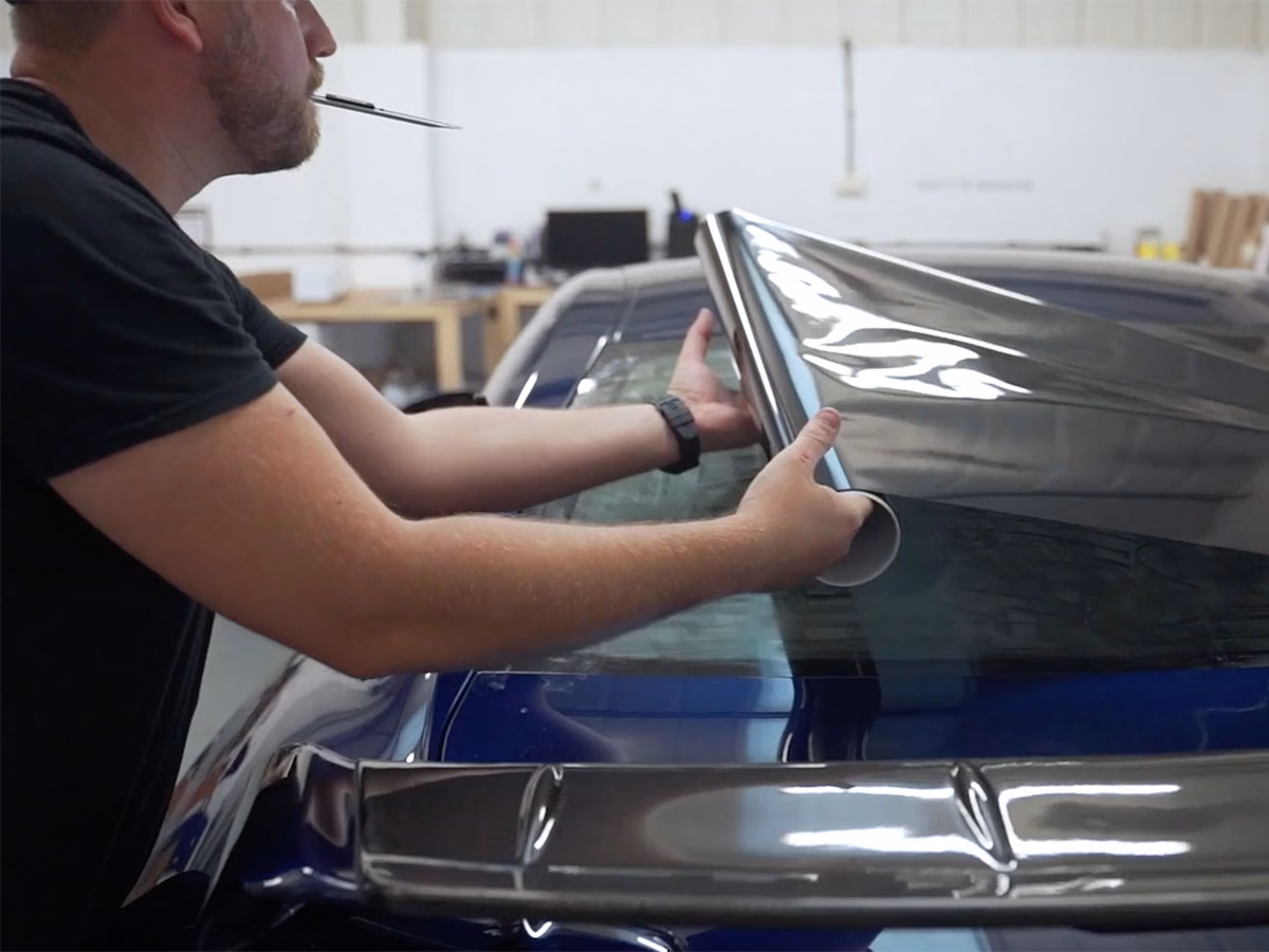 Person applying a reflective window tint film to the rear window of a blue car in a workshop